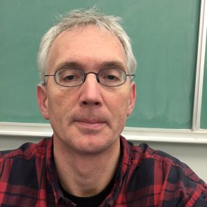 Pictured is Sean Madorin, a silver-haired man with wire-rimmed glasses wearing a flannel shirt, standing in front of a green chalkboard